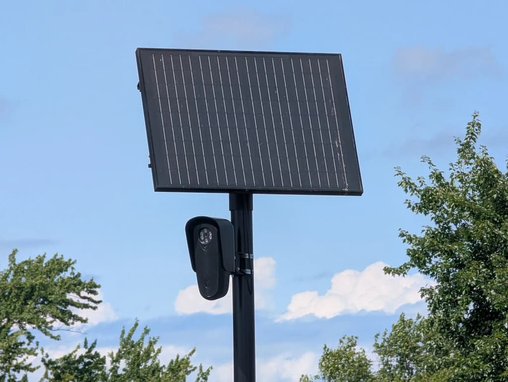 A black Flock Safety ALPR, mounted on a black pole with a black solar panel, faces left against a blue sky with trees. Photographed in a Home Depot parking lot