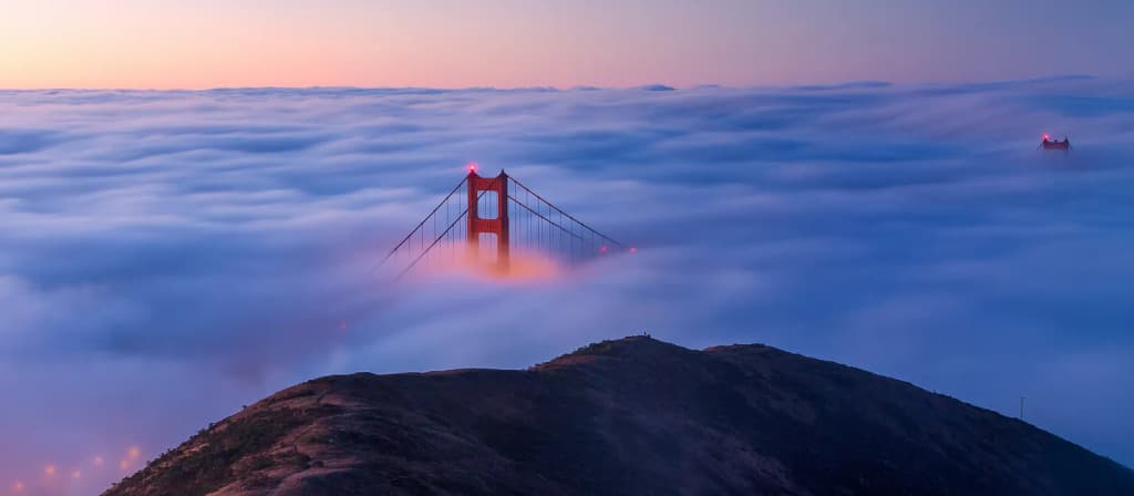 The Golden Gate Bridge as seen from the Marin Headlands at sunrise. Large parts of the bridge are covered by low, dense fog that is a frequent feature of this p